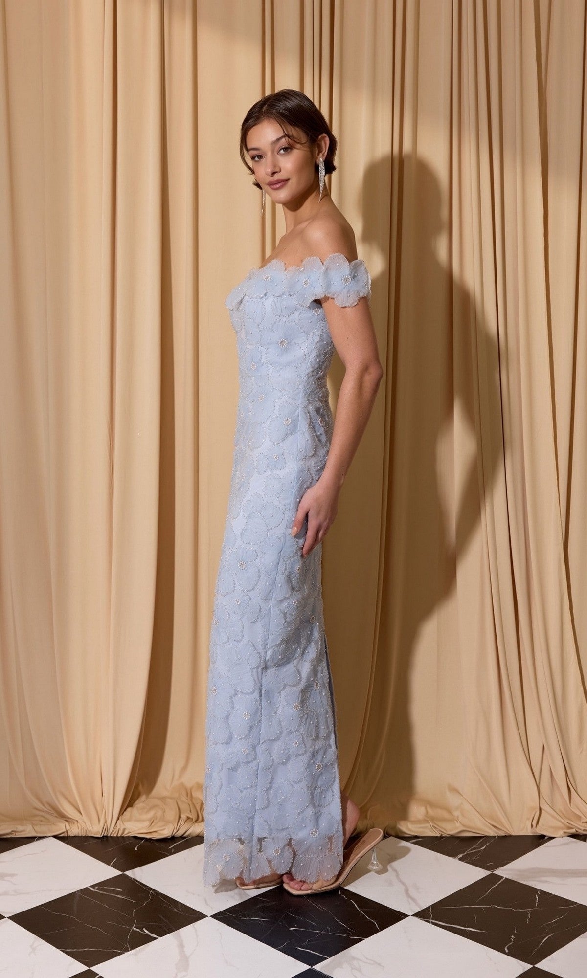 A woman stands on a black and white tiled floor, wearing the Eureka EK205 Long Prom Dress in elegant light blue lace, in front of beige curtains.